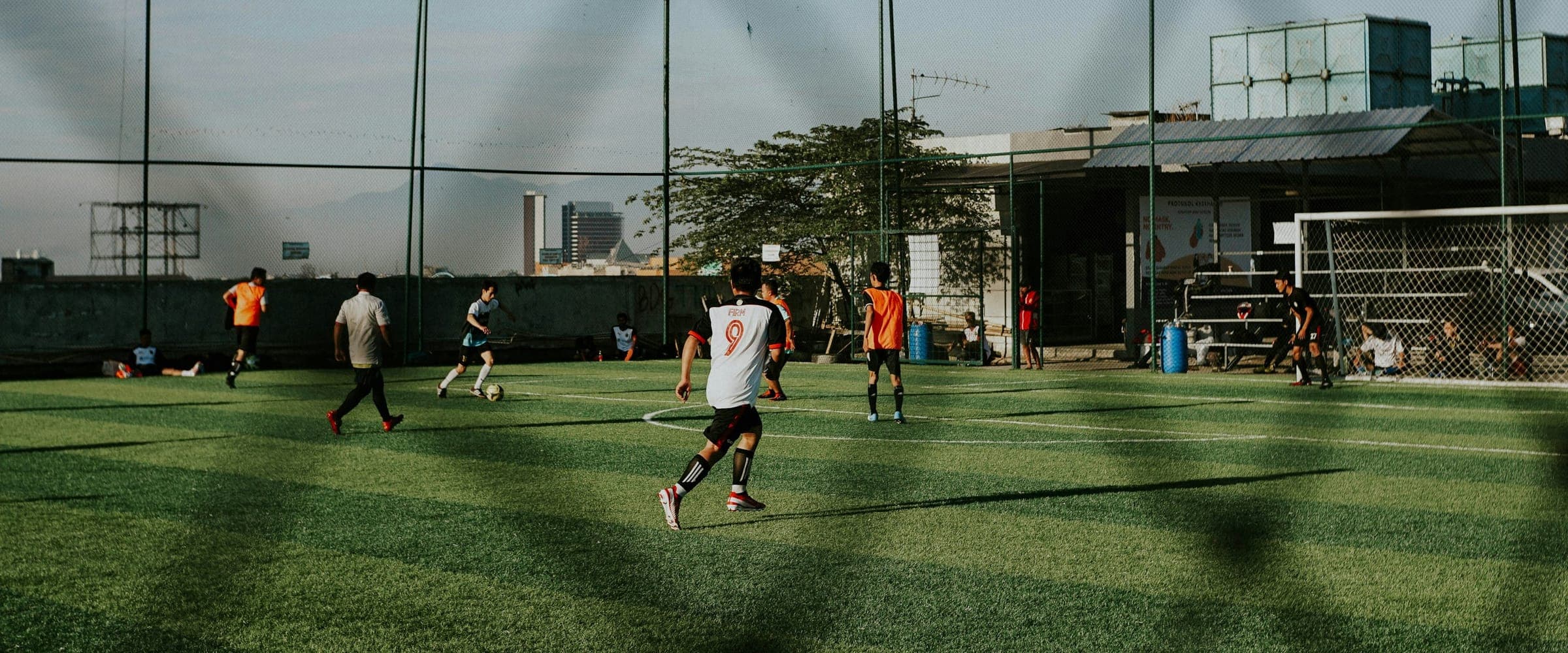 Treino de futebol society em campo de grama sintética ao entardecer, visto de fora do alambrado