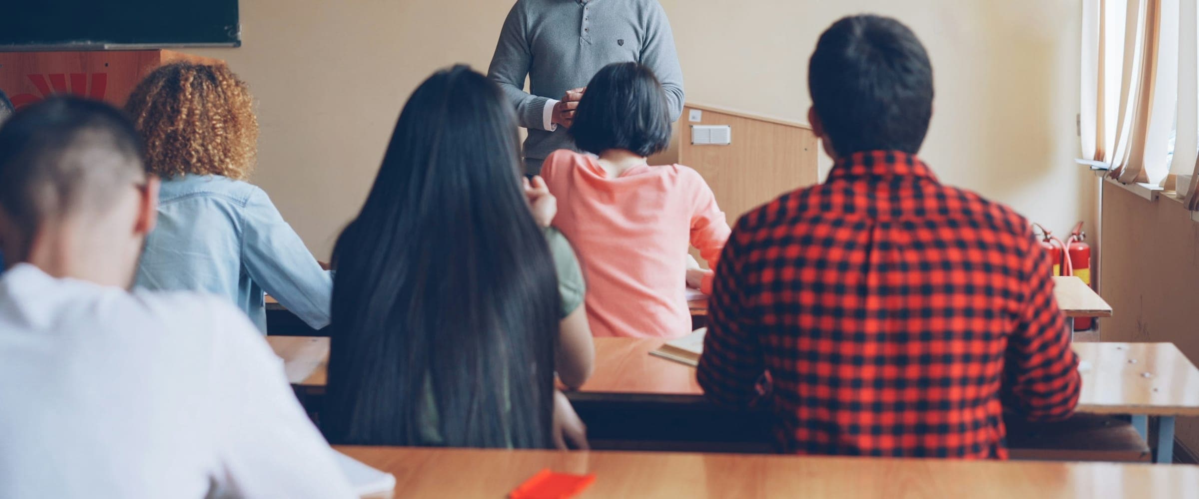 Professor em pé conversando com uma turma pequena, alunos de costas em sala clara